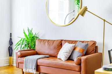 Sunlit modern living room with cognac leather sofa, patterned pillows, knit throw, gold floor lamp and round mirror, plus a green potted plant.