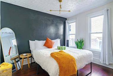 Cozy modern bedroom in an urban apartment with a dark charcoal accent wall, white fluffy bed, orange throw and pillow, wicker side table, full‑length mirror, snake plant by double windows, and hardwood floors.