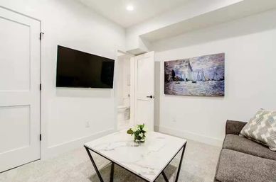 Bright modern living room with wall-mounted TV, marble-top coffee table with a small plant, gray sofa with patterned pillow, and sailboat artwork on white walls.