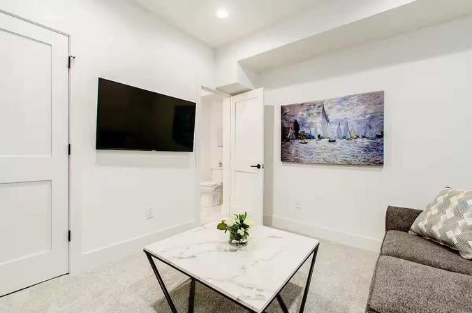 Bright modern living room interior with white walls, wall-mounted flat-screen TV, marble-top coffee table with small plant, cozy gray sofa with patterned pillow, sailboat canvas art on the wall and an open door showing a bathroom.