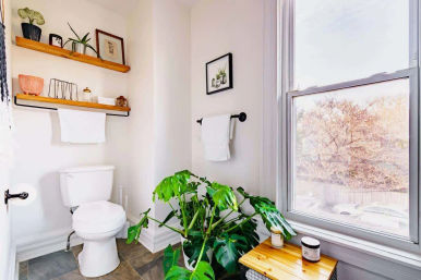 Bright, light-filled bathroom with a white toilet, wooden floating shelves, towel rails, a large window with tree view, and a lush monstera plant on tiled floor.