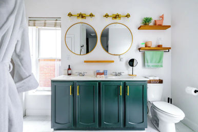 Bright modern bathroom with deep green double-sink vanity, gold hardware and light fixtures, two round mirrors above a white marble countertop, wooden floating shelves with decor, towel and toilet