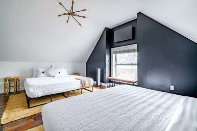 Airy attic-style bedroom with two white beds, black angled accent wall, hardwood floors, woven rugs, window bench and starburst chandelier.