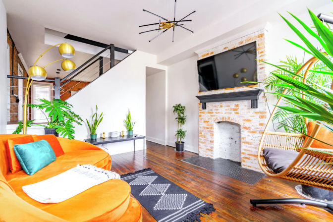 Bright modern loft living room with a curved orange velvet sofa and teal pillow, whitewashed exposed brick fireplace with wall-mounted TV, hanging rattan chair, gold arc floor lamp, indoor plants, dark hardwood floors and open metal staircase.