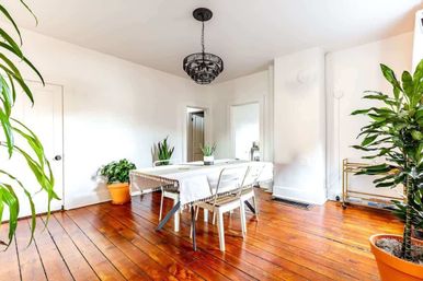 Sunlit dining room with warm hardwood floors, white walls, a rectangular table draped in a fringed cloth with metal chairs, black pendant chandelier and several potted indoor plants.