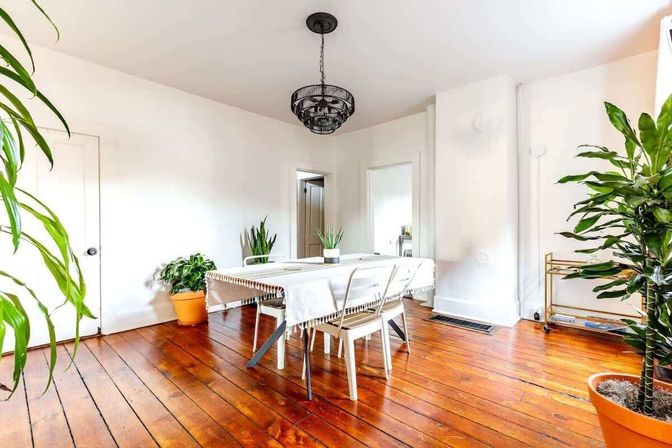 Sunlit dining room with warm hardwood floors, white walls, a rectangular table draped in a fringed cloth with metal chairs, black pendant chandelier and several potted indoor plants.
