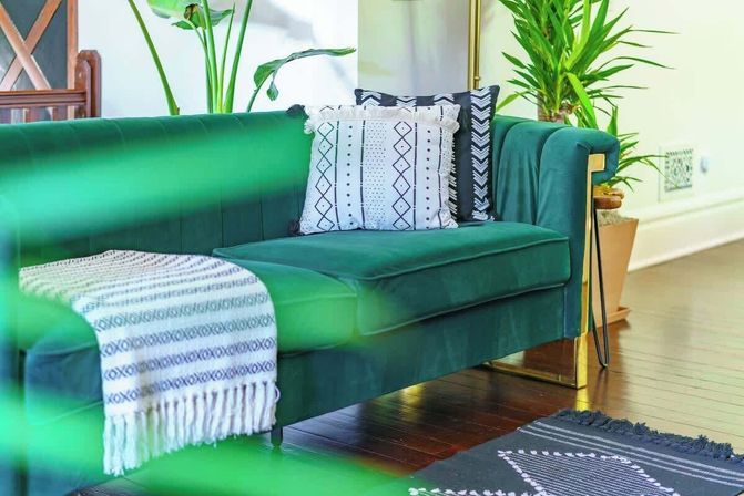 Emerald green velvet sofa in a modern living room with patterned throw blanket, textured decorative pillows, potted plants, gold-accent legs and dark hardwood floor — cozy boho-chic interior.