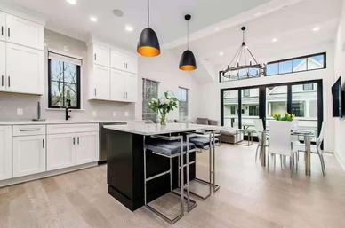 Bright, airy open-concept kitchen and dining area with white cabinetry, black island with marble countertop, chrome bar stools, pendant lights, sliding glass doors and light hardwood floors.
