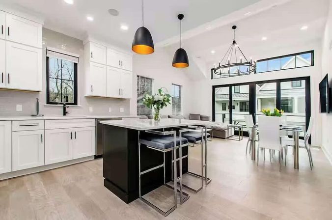 Bright, airy open-concept kitchen and dining area with white cabinetry, black island with marble countertop, chrome bar stools, pendant lights, sliding glass doors and light hardwood floors.