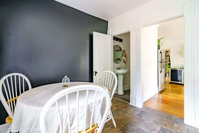 Cozy dining nook with a round table draped in white and white spindle-back chairs against a charcoal accent wall; open doors reveal a small bathroom with a pedestal sink and a bright kitchen with stainless fridge and hardwood floors.