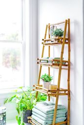 Sunlit bamboo leaning shelf beside a window holding potted plants, stacked mint-green towels and a small candle — airy indoor bathroom or spa corner.
