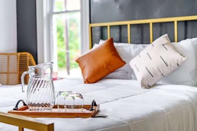 Sunlit modern bedroom with gold metal bed frame and white comforter, decorative orange and patterned pillows, and a wooden tray holding a glass pitcher and two glasses by the window.