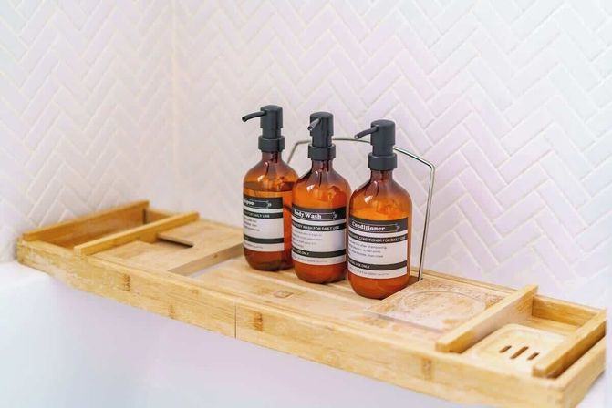 Spa-ready bamboo bathtub caddy holding three amber pump bottles labeled shampoo, body wash, and conditioner against white herringbone tile in a modern bathroom.