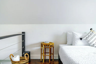 Bright minimalist bedroom corner with crisp white bedding, patterned throw pillow, bamboo side table holding an alarm clock and a woven towel basket on dark hardwood floor
