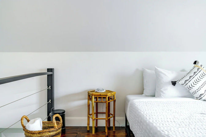 Bright minimalist bedroom corner with crisp white bedding, patterned throw pillow, bamboo side table holding an alarm clock and a woven towel basket on dark hardwood floor