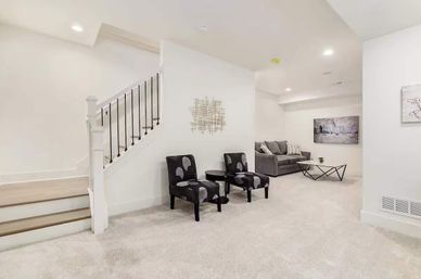 Bright finished basement lounge with carpeted floor, staircase, two patterned accent chairs and round side table, gray sofa with throw pillows, geometric coffee table and modern wall art.