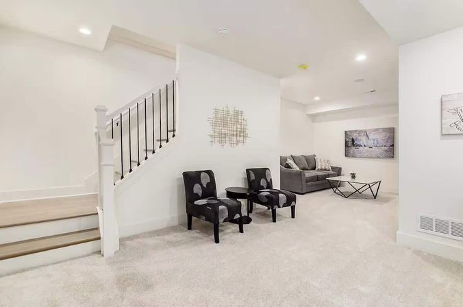 Bright finished basement lounge with carpeted floor, staircase, two patterned accent chairs and round side table, gray sofa with throw pillows, geometric coffee table and modern wall art.