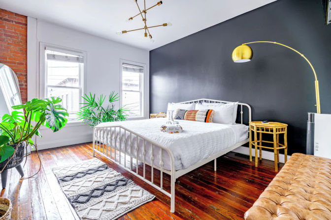 Sunlit urban loft bedroom with matte black accent wall, white metal bed on polished hardwood floors, tropical potted plants, gold arc floor lamp, rattan side table, textured rug and boho pillows.