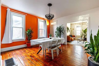 Sunlit dining room with vibrant orange accent wall, polished hardwood floors, white table with metal chairs, black hanging chandelier, potted indoor plants, and an open view into a cozy living room.