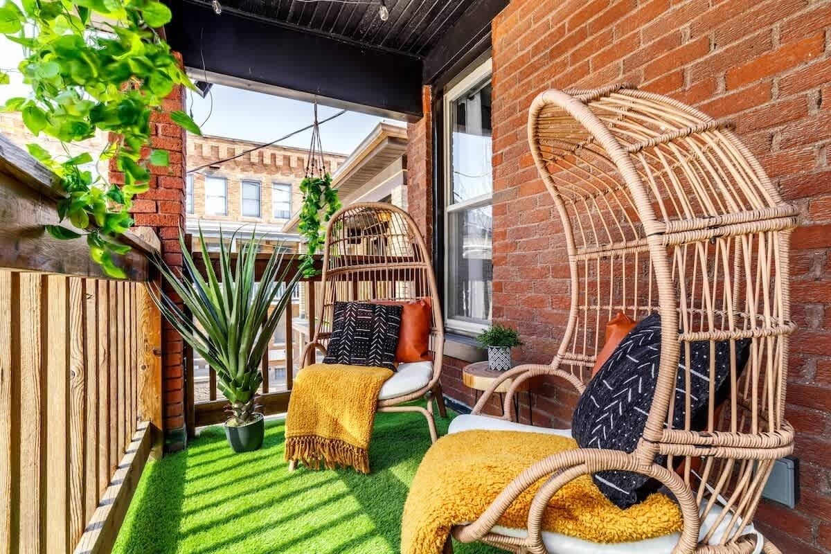 Cozy urban apartment balcony with two woven rattan egg chairs dressed in mustard throws and black patterned pillows, faux grass flooring, potted and hanging plants, and a small side table against a red brick wall.