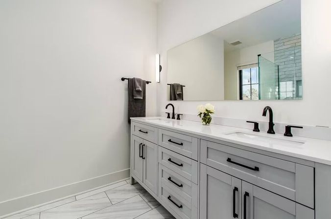 Sleek modern master bathroom with gray shaker double-sink vanity, white quartz countertop, matte black faucets and hardware, large frameless mirror, marble-look tile floor and wall towel.