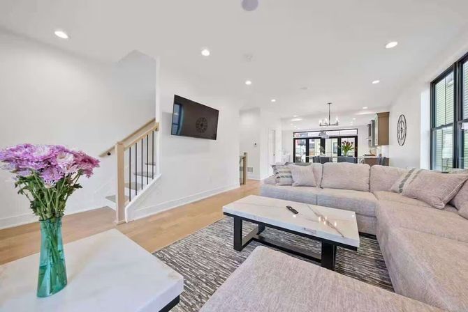 Bright contemporary open-concept living room interior with light gray L-shaped sectional, marble-top coffee table, wall-mounted TV, hardwood floors, staircase and dining area in the background, and a green vase of purple flowers in the foreground.