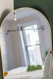 Arched gold-framed mirror in a sunlit bedroom with a playful cursive decal reading 'you're like, really pretty,' reflecting a bright window and a neatly made bed.