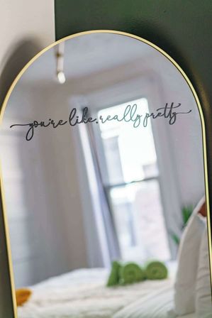 Arched gold-framed mirror in a sunlit bedroom with a playful cursive decal reading 'you're like, really pretty,' reflecting a bright window and a neatly made bed.