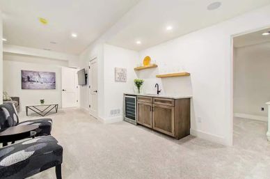 Bright modern finished basement living area with neutral white walls and carpet, recessed lighting, patterned accent chairs and coffee table, wall-mounted TV, and a wet bar with wine fridge, wood cabinets and floating shelves.