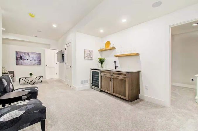 Bright modern finished basement living area with neutral white walls and carpet, recessed lighting, patterned accent chairs and coffee table, wall-mounted TV, and a wet bar with wine fridge, wood cabinets and floating shelves.