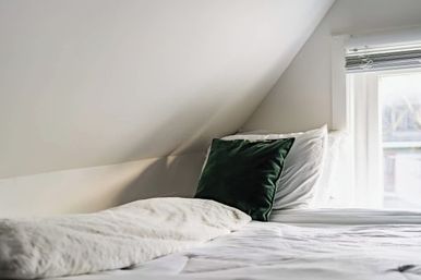Sunlit loft bedroom with sloped ceiling, crisp white bedding and a dark green velvet pillow.