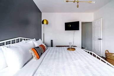 Contemporary bedroom with crisp white quilted bedding, charcoal accent wall, brass floor lamp, striped orange lumbar pillow, wall-mounted TV, woven basket and an open door.