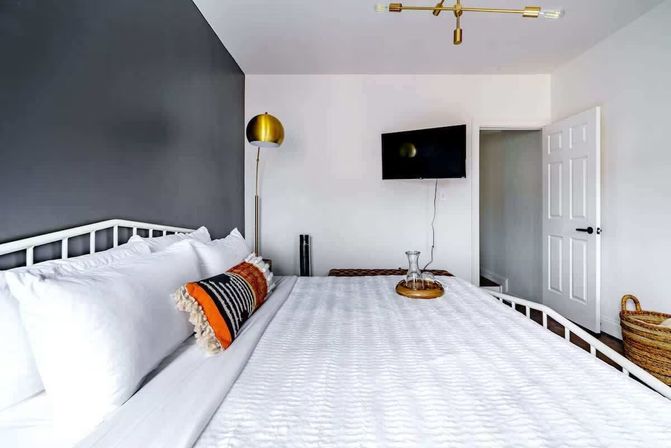 Contemporary bedroom with crisp white quilted bedding, charcoal accent wall, brass floor lamp, striped orange lumbar pillow, wall-mounted TV, woven basket and an open door.