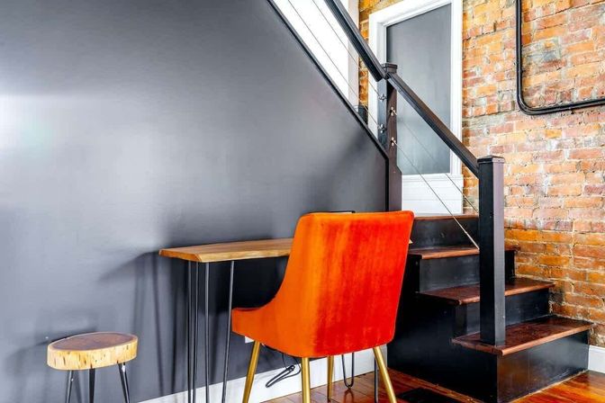 Industrial loft nook with a bright orange velvet chair at a slim wooden desk beside a black-painted staircase and exposed brick wall, small round wood stool on hardwood floor
