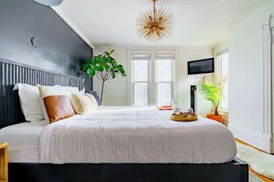 Sunlit modern bedroom interior with black accent wall, white platform bed topped with leather and textured pillows, gold starburst chandelier, indoor plants, wall-mounted TV and large windows