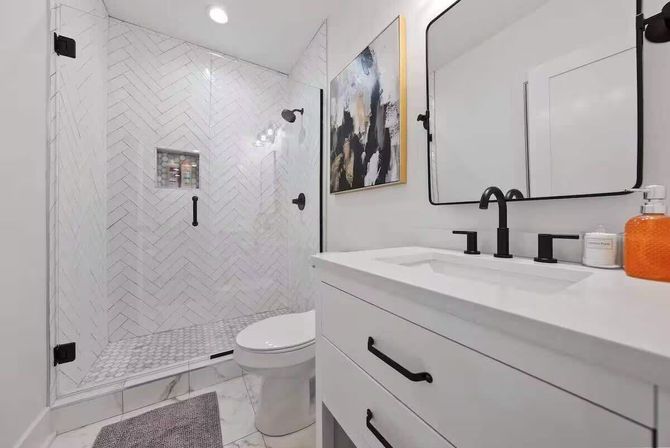 Sleek modern white bathroom with glass walk-in shower featuring herringbone marble tile, black matte fixtures, white vanity with undermount sink, large framed mirror, toilet, and bright orange soap dispenser.