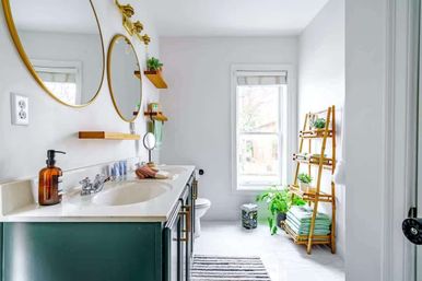 Sunlit modern bathroom with green double vanity, round gold-framed mirrors, brass sconces, wooden ladder shelf holding folded towels and potted plants.