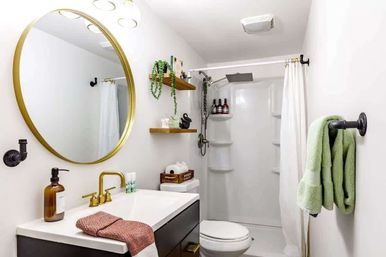 Bright modern white bathroom interior with a round gold mirror, brass faucet, black vanity, floating wood shelves with plants and decor, green towels on a hook, toilet, and a walk-in shower with rainfall showerhead and white curtain.
