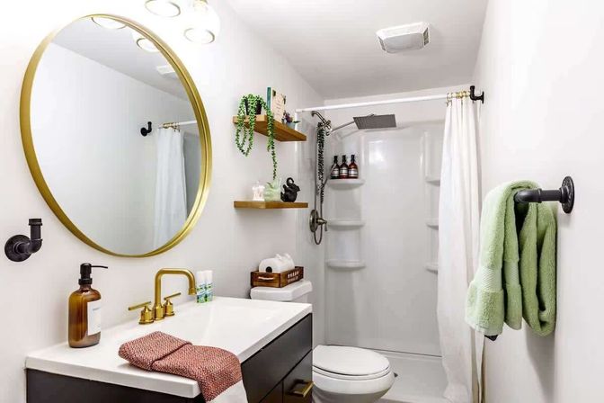 Bright modern white bathroom interior with a round gold mirror, brass faucet, black vanity, floating wood shelves with plants and decor, green towels on a hook, toilet, and a walk-in shower with rainfall showerhead and white curtain.