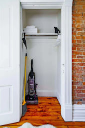 Small apartment closet with an upright vacuum and yellow-handled broom on warm hardwood floors, shelf of folded white towels and empty hangers, white trim and exposed brick wall — ready for a quick tidy.