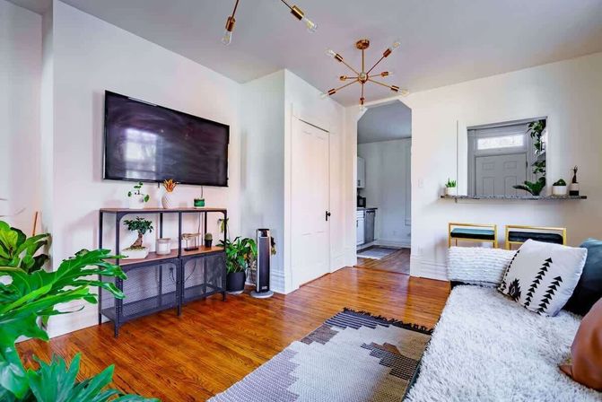 Bright modern apartment living room with wall-mounted TV, hardwood floors, potted plants, mid-century chandelier, open kitchen pass-through and cozy white sofa