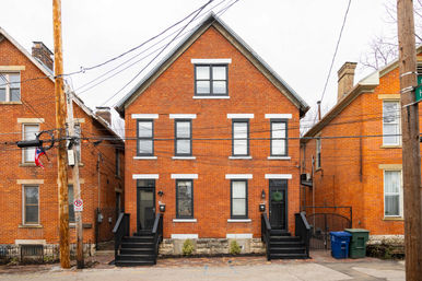 Red brick gabled duplex on an urban residential street, black-trimmed windows and twin black stoop entrances, overhead utility wires and wooden poles, blue recycling and green trash bins at the curb.