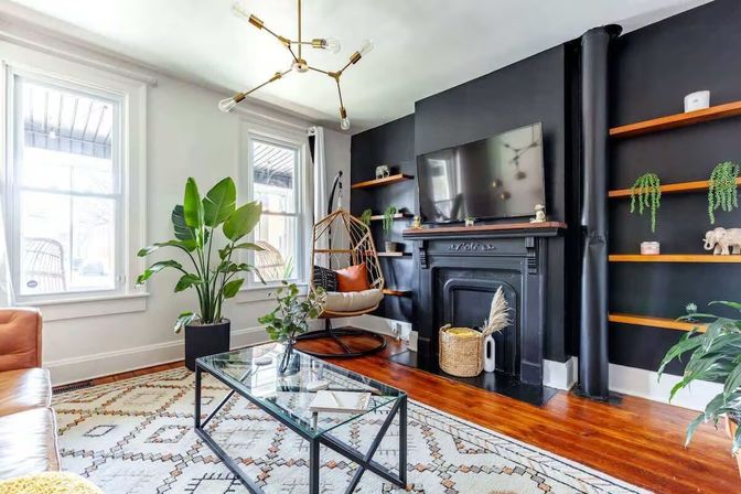 Bright modern living room with large windows, mid-century brass chandelier, black accent wall with vintage fireplace and wall-mounted TV, hanging rattan chair, oversized houseplants, glass coffee table, patterned rug and hardwood floors.
