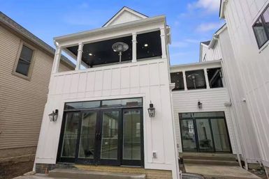 Modern white two-story townhouse facade with covered second-floor balcony and patio heaters, black framed glass doors, lantern sconces, narrow urban lot.