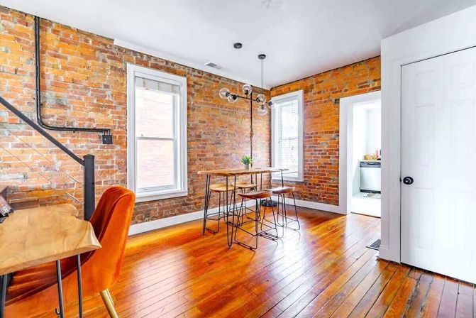 Cozy loft-style dining area with exposed brick walls, glossy hardwood floors, white-trim windows, industrial globe pendant light, wood-and-metal high table with stools, an orange velvet chair and open doorway to the kitchen.