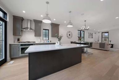 Bright open-concept modern kitchen and living room with a large dark island topped in white quartz, stainless steel range hood, gray cabinets, pendant lights and light hardwood floors