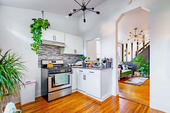 Bright open-concept modern kitchen in an urban apartment with white cabinets, stainless steel stove, mosaic tile backsplash, hanging pothos and hardwood floors leading to a green sofa in the living area.