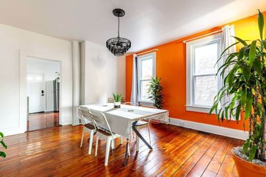Sunlit dining room with bright orange accent wall, glossy hardwood floors, white table, black chandelier and potted plants