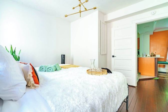 Bright modern bedroom with fluffy white comforter, teal rolled towels and orange accent pillow, brass chandelier, hardwood floors, indoor plant, and open door revealing a kitchen with an orange accent wall.