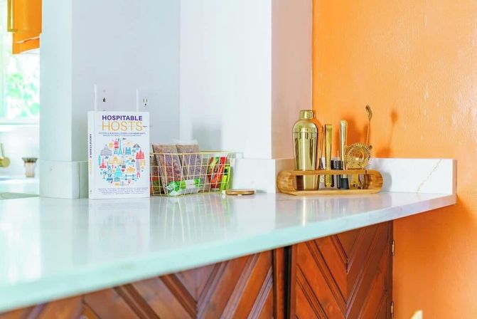 Bright kitchen countertop with white marble surface, orange accent wall and wooden chevron cabinets, featuring a cookbook titled "Hospitable Hosts", a wire snack basket and brass cocktail tools on a bamboo tray — modern home bar setup.
